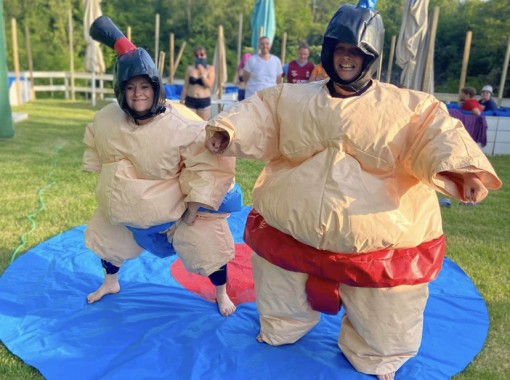 Deux femmes en tenues de sumo gonflables s’affrontent dans une ambiance ludique et amusante. Les costumes volumineux ajoutent du fun à leur duel, offrant un spectacle divertissant.